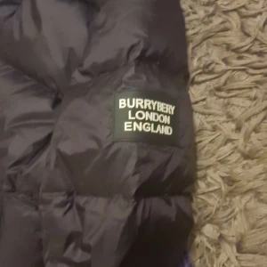 Svart pufferjacka från Burberry - Säljer en svart pufferjacka från Burberry i mycket bra skick. Jackan är perfekt för kalla dagar med sin quiltade design och långa ärmar. Den har en praktisk dragkedja och knappar framtill. Passar både höst och vinter. Storleken är inte angiven, men den ser ut att ha en normal passform.
