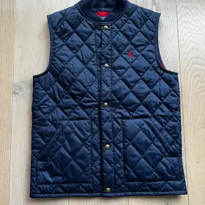 Säljer en snygg mörkblå quiltad väst från Ralph Lauren. Västen har en broderad röd logga på bröstet och stängs med knappar. Perfekt för höst och vår, och passar bra över en tröja för extra värme. Den har två praktiska fickor framtill. Storleken är 14-16 års ålder som motsvarar XS-S.