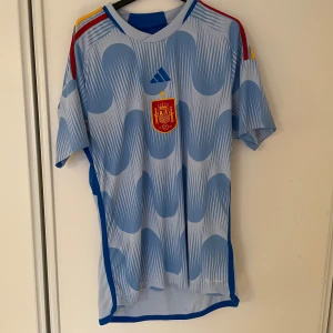 Spanien fotbollströja från 22/23 - Säljer en fotbollströja  i storlek L. Tröjan har korta ärmar och ett vågigt mönster i olika blå nyanser. Några små fläckar finns på baksidan. Perfekt för fotbollsfans!