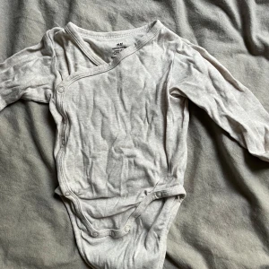 Beige omlottbody från H&M - Säljer en söt och bekväm beige omlottbody från H&M i ekologisk bomull. Perfekt för de allra minsta med praktiska tryckknappar för enkel på- och avklädning. Långärmad och mjuk mot huden, idealisk för daglig användning.