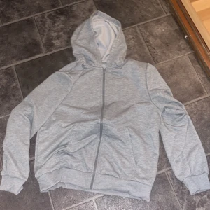 Grå hoodie med dragkedja - Säljer en stilren grå hoodie i mycket bra skick. Den har en praktisk dragkedja framtill och en skön huva. Perfekt för både höst och vår, och funkar lika bra till träning som till vardags. Storleken är normal och materialet känns mjukt och bekvämt.