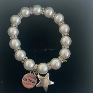 Pärlarmband med berlocker - Snyggt pärlarmband med vita pärlor och glittriga detaljer. Armbandet har två berlocker, en rund med texten 'Isadora' och en stjärnformad. Perfekt för att ge en elegant touch till din outfit. Passar både till vardag och fest!