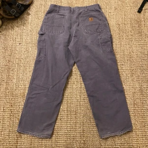 Grålila byxor från Carhartt - Säljer ett par grå Carhartt byxor i Dungaree Fit. De är tillverkade i 100% bomull och har en avslappnad passform med flera praktiska fickor. Perfekta för vardagsbruk eller arbete. Byxorna är i bra skick och redo för nya äventyr!