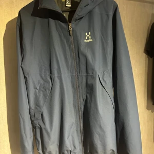 Blå vindjacka från Haglöfs - Säljer en snygg blå vindjacka från Haglöfs i mycket bra skick. Jackan har en dragkedja framtill och är perfekt för höst och vår. Den är långärmad och har en praktisk huva. Perfekt för utomhusaktiviteter och vardagsbruk!