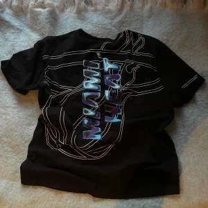 Svart Miami Heat t-shirt från NBA - Säljer en svart t-shirt med Miami Heat-tryck från NBA. Den har en cool design med texten 'Miami Heat' i blått och vitt. Perfekt för basketfans! T-shirten är i bra skick och passar perfekt till vardags eller sportiga tillfällen.