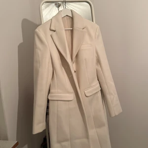 Vit kappa från ASOS - Säljer en elegant lång, off white - kappa i mycket bra skick. Den har en klassisk design med långa ärmar och knappar framtill. Perfekt för höst och vinter, och ger en stilren look till vilken outfit som helst. Passar både till vardags och festliga tillfällen. Aldrig använts förut ( testades 1x vid inkomst av beställning). 