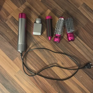 Multifunktionellt hårstylingverktyg med tillbehör - Säljer ett mångsidigt hårstylingverktyg med flera tillbehör för olika stylingbehov. Inkluderar en huvuddel med sladd och fyra utbytbara tillbehör: en hårtork, en locktång, en volymborste och en rundborste. Perfekt för att skapa olika frisyrer med lätthet. Verktyget har en snygg design i silver och rosa.