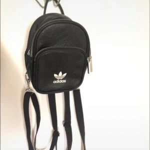 Svart mini ryggsäck från Adidas - Säljer en liten och smidig svart ryggsäck från Adidas. Den är i svart läder med en snygg textur och har det klassiska Adidas-loggan på framsidan. Ryggsäcken har justerbara axelband och en dragkedja för säker förvaring. 