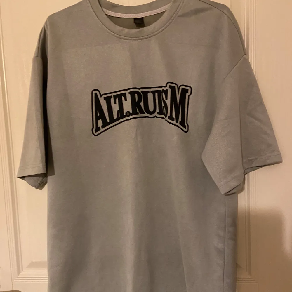 Säljer en grå t-shirt med ett coolt svart tryck 'Altruism' på framsidan. T-shirten är i normal passform och har korta ärmar. Perfekt för vardagsbruk eller en avslappnad dag ute. Materialet känns mjukt och bekvämt. Passar bra till jeans eller shorts!. T-shirts.