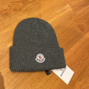 Beanie mössa Grå stickad mössa Moncler liknande helt ny  - Säljer en snygg Beanie mössa i grått. Den är Moncler liknande i modell och stil. Den är i nytt skick och har en broderad logga framtill. Perfekt för kalla dagar och ger en stilren look. Mössan är mjuk och bekväm, idealisk för både höst och vinter.