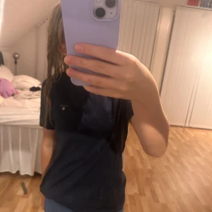 Svart t-shirt från GANT - Säljer en stilren mörkblå t-shirt från GANT i juniorstorlek men passar mig som har XS i vanliga fall. Den har en liten logga på bröstet och är i mycket bra skick. Perfekt för vardagsbruk eller en avslappnad dag ute. Passar bra till jeans eller shorts!