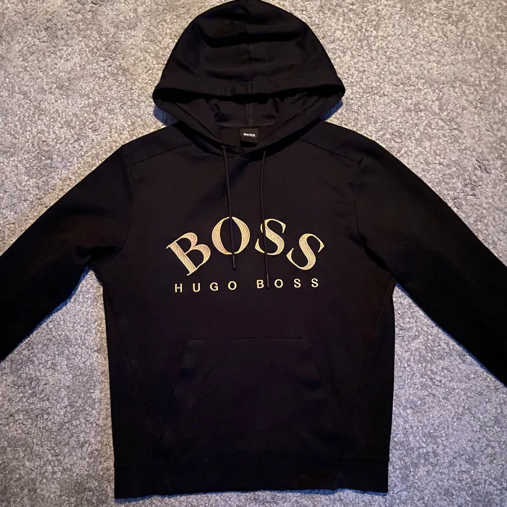 Säljer en snygg svart hoodie från Hugo Boss med stort BOSS-tryck i guld på framsidan, perfekt för en stilren och bekväm look. Lite små defekter men annars en skön tröja! . Tröjor & Koftor.