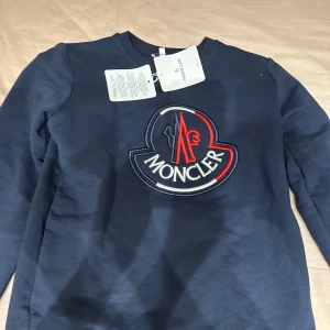 Mörkblå tröja från Moncler - Säljer en snygg mörkblå tröja från Moncler med deras ikoniska logga broderad på framsidan. Tröjan har långa ärmar och är perfekt för både höst och vinter. Den är i mycket bra skick och passar perfekt för en stilren look.