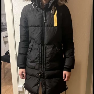Svart dunjacka från Parajumpers - Säljer en svart dunjacka från Parajumpers i mycket bra skick. Jackan är lång och har en snygg pälskantad huva. Perfekt för kalla vinterdagar med sin varma och skyddande design. Den har en dragkedja och knappar framtill samt praktiska fickor. En stilren och funktionell jacka för vintern! Kvitto finns inte men det finns en QR kod som man kan skanna för at kunna se om den är äkta 