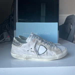 Philippe Model sneakers i vit - Säljer ett par Philippe Model sneakers i vit mocka med coola slitningar och vita detaljer. Skorna har en snygg glittrig häl och klassisk snörning. Perfekta för en avslappnad stil med en touch av lyx. Sulan har tappat färgen lite. De är i Okej skick och kommer med originalkartong. Dom är såklart äkta!