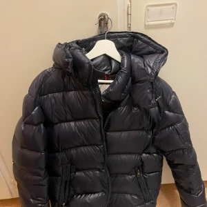 Marinblå pufferjacka från Moncler - Säljer en snygg pufferjacka från Moncler i mycket bra skick. Den har ett litet fel som jag vet om (skriv pm). Annars är den fett skön och varm med fungerande NFC chip i Moncler loggan. Säljer eftersom det inte riktigt är min stil och vill ha en annan jacka. Storlek 5 så den borde passa M och L (är 184 och den passar bra). Pris kan diskuteras. OBS!! replika.