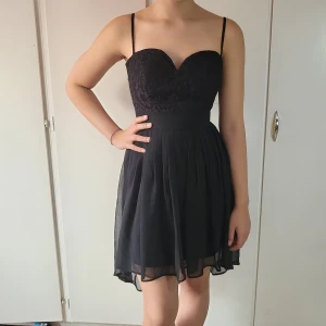 Klänning - Svart klänning i chiffong med vadderad spets vid bysten och dragkedja i rygg. Kjoldelen kortare fram och längre bak. Ny/oanvänd (prislapp kvar)