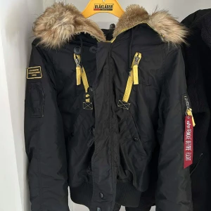 Svart jacka med päls från Alpha Industries - Säljer en snygg svart jacka från Alpha Industries i storlek S. Den har en mysig pälsfodrad huva och gula detaljer som ger en cool kontrast. Jackan har dragkedja och knappar framtill, samt flera fickor för praktisk förvaring. Perfekt för höst och vinter!