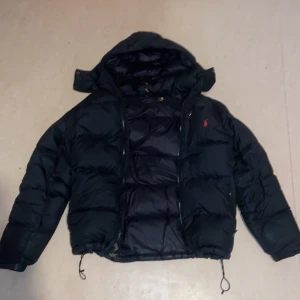 Svart polo pufferjacka  - Säljer en svart pufferjacka i bra skick. Den är perfekt för kalla vinterdagar med sin tjocka vaddering. Jackan har en dragkedja framtill och justerbara snören i nederkanten för extra skydd mot kylan. Den har också en liten röd logga på bröstet som ger en snygg detalj.