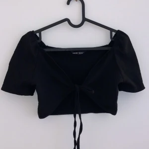 Svart kortärmad topp med knyt - Snygg svart kortärmad topp från H&M med knyt framtill. Perfekt för en somrig dag eller en utekväll. Toppen är croppad och har en stilren design som passar till många olika outfits.
