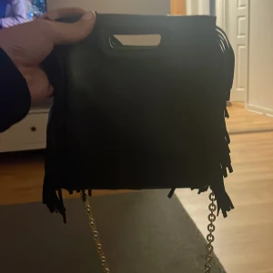Svart axelväska med fransar - Säljer en snygg svart axelväska med fransar och kedjedetaljer. Perfekt för en kväll ute eller en dag på stan. Väskan har ett handtag upptill och är i mycket bra skick. Den är både stilren och trendig!