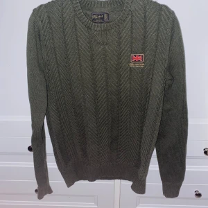 Grön stickad tröja från Morris - Säljer en snygg grön stickad tröja från Morris. Den har ett diskret märke med brittisk flagga på bröstet och ett klassiskt kabelstickat mönster. Perfekt för kyliga dagar och passar både till vardags och lite finare tillfällen. Tröjan har långa ärmar och en rund halsringning.