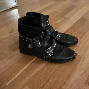 Svarta boots med nitar och spännen - Säljer ett par svarta boots i skinn med coola nitar och spännen. De har en rockig stil och är perfekta för höst och vinter. Skorna har en spetsig tå och ser ut att vara i bra skick. Perfekta för att ge din outfit en edgy touch!