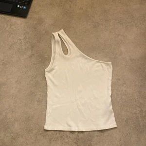 Vit oneshoulder topp från BikBok - Säljer en stilren vit oneshoulder topp från BikBok i storlek M. Perfekt för sommardagar eller en utekväll. Toppen är i ribbat material och har en tight passform. Superfin att matcha med jeans eller kjol!