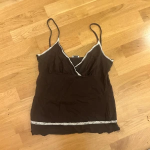 Brun linne med spetsdetaljer - Säljer ett brunt linne med smala axelband och v-ringning. Det har fina vita spetsdetaljer längs kanterna och en dekorativ spetskant nedtill. Perfekt för sommardagar eller som ett lager under en kofta. Den är från intimissimi❤️