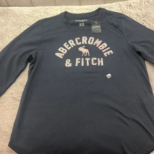 Mörkblå tröja från Abercrombie & Fitch - Säljer en snygg mörkblå tröja från Abercrombie & Fitch i storlek 13/14. Den har ett glittrigt tryck med märkesnamnet och en älg på framsidan. Perfekt för en casual look eller skoldagar. Tröjan är i nytt skick och superbekväm! Den är i ny skick med prislappen kvar.