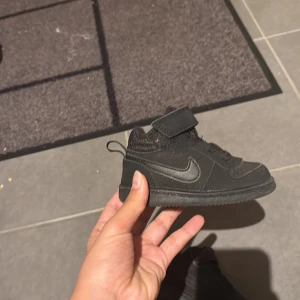 Svarta Nike sneakers för barn/babys - Säljer ett par svarta Nike sneakers för babysar mycket bra skick. Skorna har en stilren design med kardborreband och snörning för enkel på- och avtagning. Perfekta för både lek och vardag!