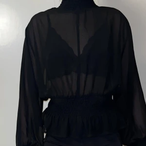 Svart transparent blus - Snygg svart transparent blus med långa ärmar och smockdetaljer i midjan. Perfekt för en kväll ute eller en fest. Den har en elegant och luftig design som ger en chic look. Passar bra över ett linne eller en snygg bh.