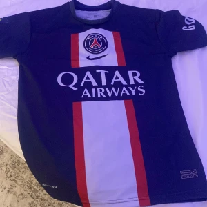 Blå PSG fotbollströja - Säljer en blå PSG fotbollströja från Nike i Dri-FIT material. Tröjan har korta ärmar och ett snyggt rött och vitt mönster på framsidan med Qatar Airways som sponsor. Perfekt för alla PSG-fans! 🏆
