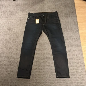 Mörkblåa jeans från G-Star RAW - Säljer ett par snygga mörkblåa jeans från G-Star RAW i modell 3301. De är i nytt skick med etikett kvar. Jeansen har en slim passform och är tillverkade i ett slitstarkt denimtyg. Perfekta för både vardag och fest!