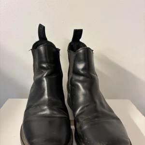Svarta Chelsea boots - Säljer ett par svarta Chelsea boots i skinn. De har en klassisk design med elastiska paneler på sidorna för enkel på- och avtagning. Perfekta för både vardag och fest. Skorna är i bra skick med lite slitage som ger dem en cool look. Billiga! 