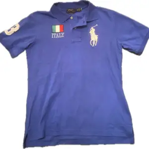 Säljer en snygg blå polo från Ralph Lauren med broderade detaljer. Den har korta ärmar och nummer 3 på ärmen och ryggen. Italienska och amerikanska flaggor pryder framsidan och baksidan. Perfekt för en sportig look!