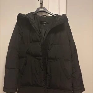 Svart dunjacka med huva - Säljer en svart dunjacka från vero moda i mycket bra skick. Jackan har en praktisk huva och dragkedja framtill. Perfekt för kalla vinterdagar med sin varma och vadderade design. Den har långa ärmar och en stilren look som passar till det mesta. Perfekt för både vardag och lite finare tillfällen.