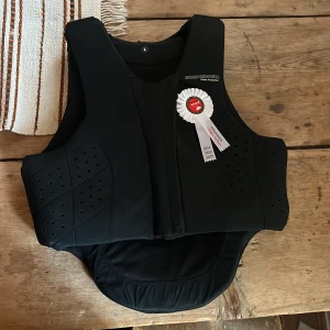 Svart säkerhetsväst från Komperdell - Säljer en svart säkerhetsväst från Komperdell, perfekt för ridning. Västen har en frontdragkedja och är CE-godkänd med Level 3 skydd enligt EN 13158 standard. Den är i nyskick och har en snygg, modern design. Passar perfekt för dig som vill ha både stil och säkerhet när du rider. Aldrig använd, bara provad. Köpt våren 2023 men pga skada hann den aldrig användas. Nypris 2995 kr.