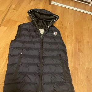 Svart dunväst från Moncler - Säljer en snygg svart dunväst från Moncler i mycket bra skick. Västen har en dragkedja framtill och en broderad Moncler-logga på bröstet. Den är perfekt för höst och vår, med en stilren design och en praktisk huva. Passar både till vardags och lite finare tillfällen.