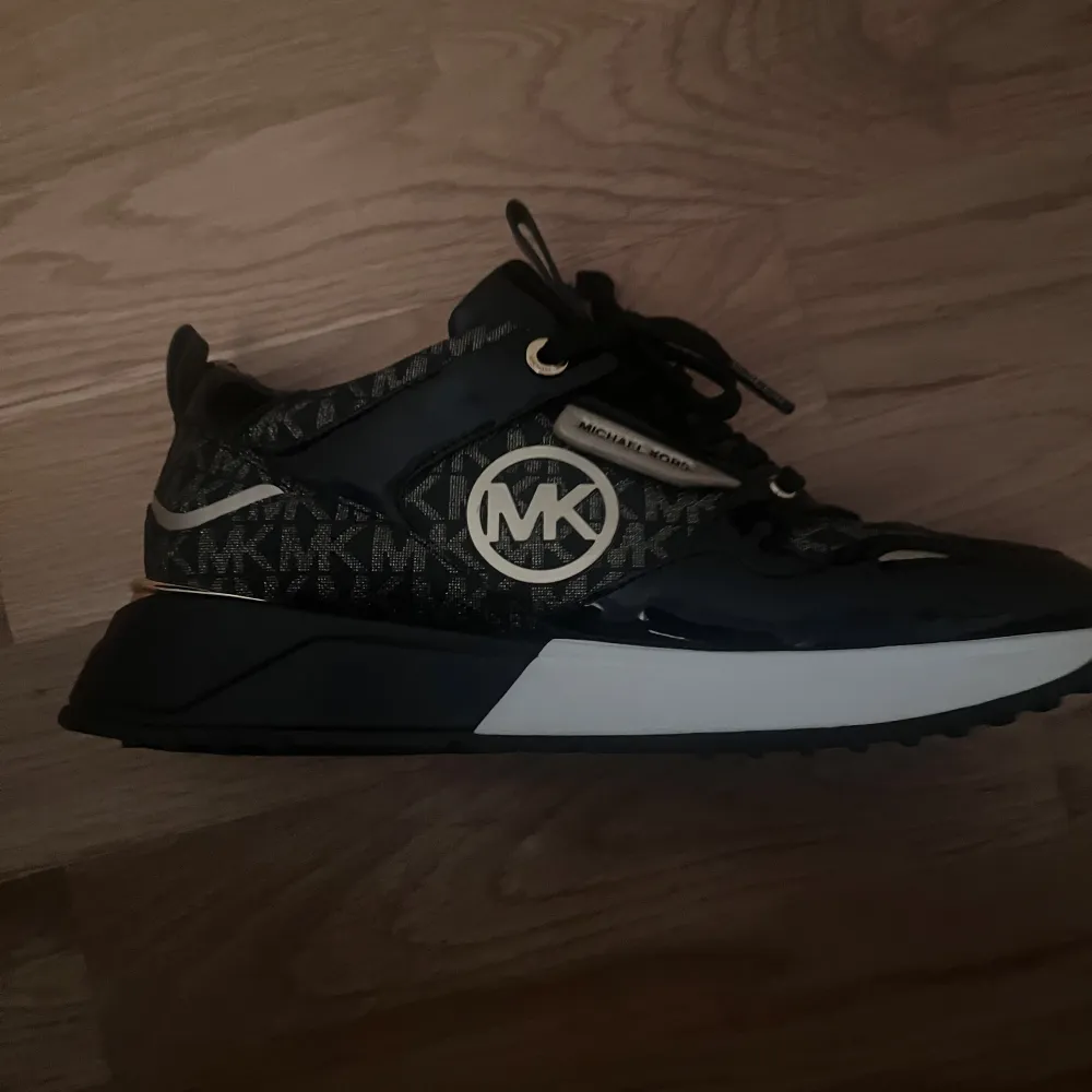 Snygga svarta sneakers från Michael Kors med gulddetaljer och logotyp. Skorna har ett mönster med märkets initialer och är i mycket bra skick. Perfekta för både vardag och festliga tillfällen. De har en bekväm passform och snörning framtill. Storlek 38 ord pris 2300 kr. Pris kan diskuteras!. Kengät.
