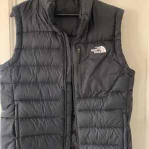Svart dunväst från The North Face str M - Säljer en svart dunväst från The North Face i mycket bra skick. Västen har en dragkedja framtill och är perfekt för höst och vinter. Den är varm och stilren, idealisk för både vardag och utomhusaktiviteter. (Nyskick). Str Medium