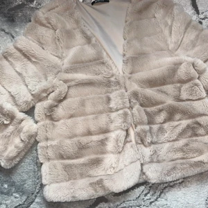 Beige pälsjacka - Supermjuk och fluffig beige jacka som är perfekt för att hålla sig varm och stilren under kyliga dagar. Jackan har en lyxig känsla med sitt täta pälsmaterial och är designad för att ge en elegant look. Perfekt för höst och vinter!