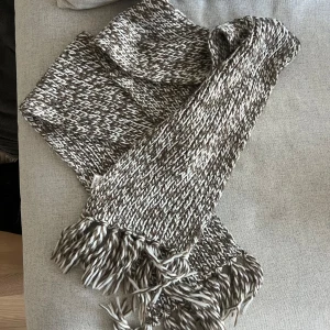 Stickad halsduk med fransar - Säljer stickad halsduk i brun/grått och vitt. Den är snabba använd och i mycket bra skick. Lagom lång! 