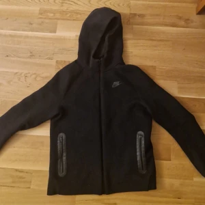 Svart nike tech dres - Säljer en svart hoodie från Nike i storlek xs .barn Den har en dragkedja framtill och en klassisk  Nike  på bröstet. Perfekt för höst och vinter, och passar både till träning och vardagsbruk. Superbekväm och stilren design! Kan mötas Stockholm Skogås elr Skärholmen 
