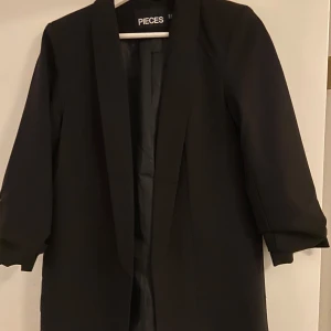Svart kavaj från Pieces - Snygg svart kavaj från Pieces i bra skick. Perfekt för både vardag och fest! Den har en klassisk design med trekvartsärmar och en stilren passform. Passar utmärkt till både jeans och klänning.