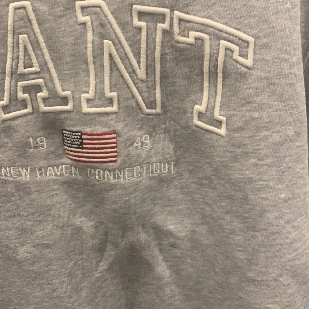Säljer en snygg grå sweatshirt från GANT i bra skick. Den har ett klassiskt tryck med GANT-loggan och en liten amerikansk flagga på framsidan. Perfekt för en avslappnad stil och passar bra till både jeans och joggers. Skön och stilren för vardagsbruk!. Neuletakit & Villapaidat.