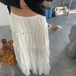 Vit långkjol med volanger - Superfin vit långkjol i bomull med volanger. Perfekt för sommardagar eller en avslappnad vårlook. Kjolen har ett elastiskt midjeband för en bekväm passform. Den broderade designen ger en romantisk touch. Passar perfekt till en enkel topp eller blus.