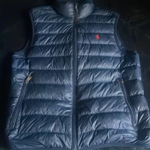 Blå dunväst från Ralph Lauren - Säljer en snygg blå dunväst från Ralph Lauren med dragkedja och röd logga på bröstet. Perfekt för höst och vår, håller dig varm och stilren. Västen har två praktiska fickor med dragkedja. Passar bra till både vardag och fest! Storlek S och i utmärkt skick. Hör av dig!