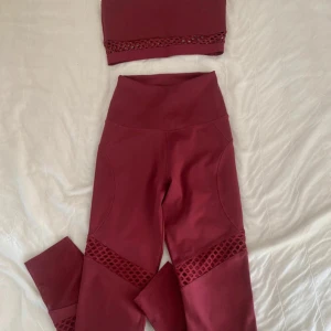 Vinrött träningsset med meshdetaljer - Snyggt vinrött träningsset från Gymshark med en croppad topp och matchande leggings. Båda delarna har coola meshdetaljer som ger en extra touch. Perfekt för gymmet eller yoga! Setet är i ett mjukt och stretchigt material som ger bra passform och komfort. Knappt använda.
