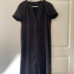 Svart långklänning med knappar - Säljer en stilren svart långklänning i storlek Xs. Klänningen har korta ärmar och knappar hela vägen ner framtill. Perfekt för både vardag och festliga tillfällen. Enkelt att matcha med olika accessoarer för att skapa din egen stil!